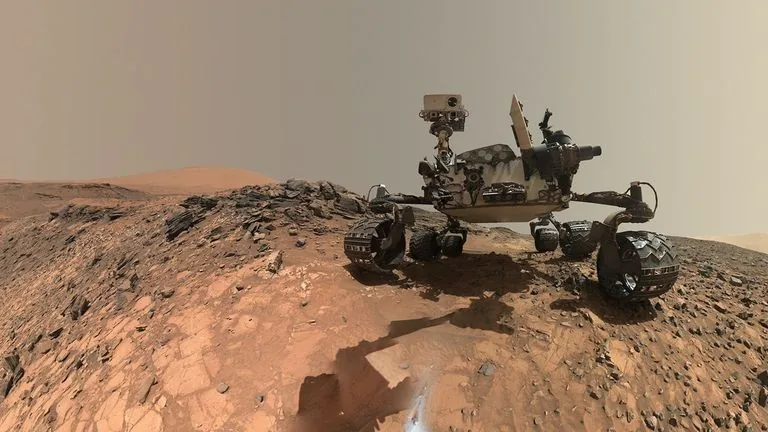 Un rover sobre la superficie marciana ondulada y anaranjada, bajo un cielo gris y plano, rodeado de trozos de roca.