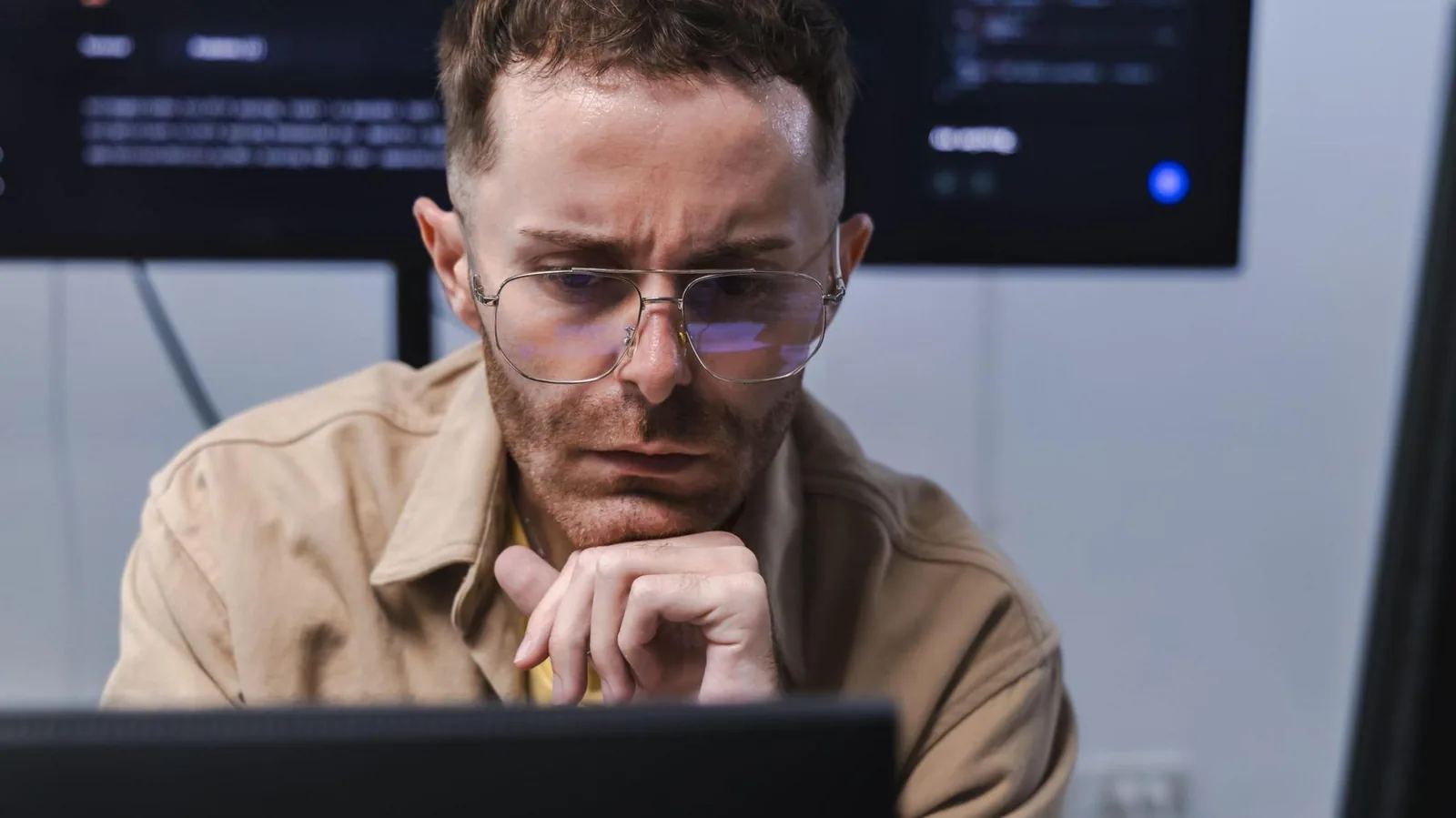 Un ingeniero de software con camisa café claro mira preocupado un monitor, con una pantalla grande con código en la pared detrás.